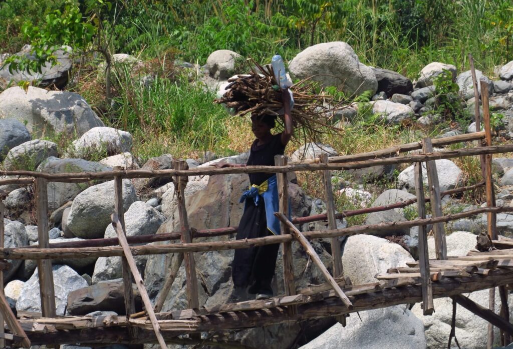 Frau balanciert Holzbündel auf ihrem Kopf über eine wackelige Holzbrücke in den Rwenzori Bergen